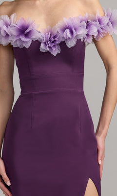 A person wearing the Mac Duggal 21235 long formal dress, which features an off-the-shoulder sweetheart neckline, a fitted waist, a thigh-high slit, and decorative lavender flowers along the neckline in purple crepe-jersey.
