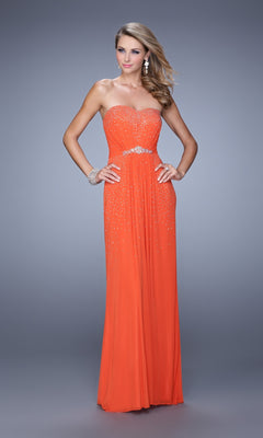 A woman models the La Femme Embellished-Jersey Prom Dress 21237, a floor-length orange strapless gown with silver accents and jeweled belt, paired with matching jewelry, set against a plain gray background.