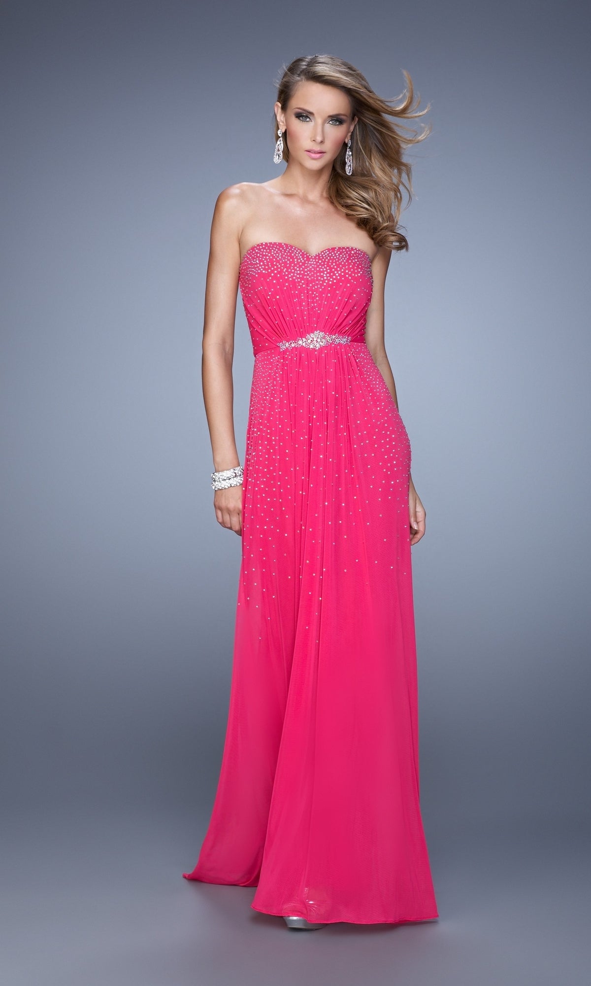 A woman in a strapless, floor-length La Femme Embellished-Jersey Prom Dress 21237 in bright pink with beaded accents and a silver belt stands against a plain grey background, accessorized with matching silver jewelry.