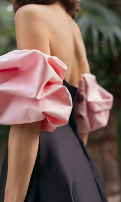 A woman stands outdoors with blurred greenery in the background, wearing the Long Formal Dress: Mac Duggal 21241—a black strapless satin gown featuring large pink satin puff sleeves.
