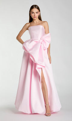A woman wearing the Mac Duggal 21245 long formal dress in ice pink, featuring a strapless design, floor-length silhouette, large bow detail at the waist, and a thigh-high slit, stands against a plain background.