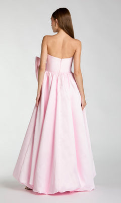 A woman with long straight hair stands with her back to the camera against a plain background, wearing the Mac Duggal 21245—a stunning, floor-length pastel pink formal dress with a strapless neckline and full skirt.