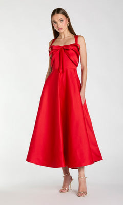 A woman standing in a sleeveless, tea-length red Mac Duggal 21247 party dress with a large bow detail on the bodice, paired with gold high-heeled sandals.