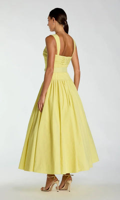 A woman stands in profile with her back to the camera, wearing the Tea-Length Party Dress: Mac Duggal 21251—a pale yellow, sleeveless, mid-length dress featuring a fitted bodice and flared skirt—paired with nude high-heeled sandals.