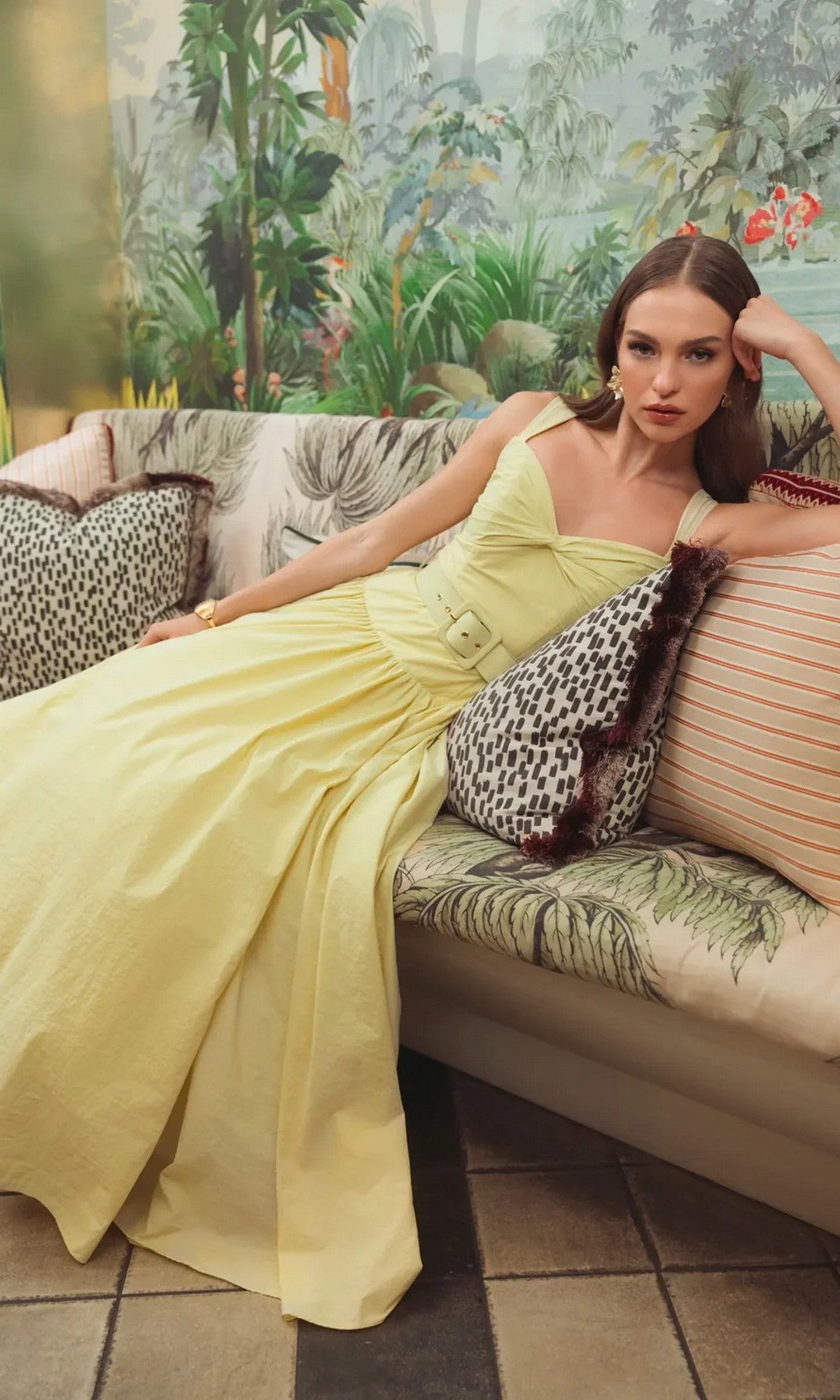 A woman in a tea-length Mac Duggal 21251 party dress sits on a patterned sofa, with tropical-themed wallpaper in the background.