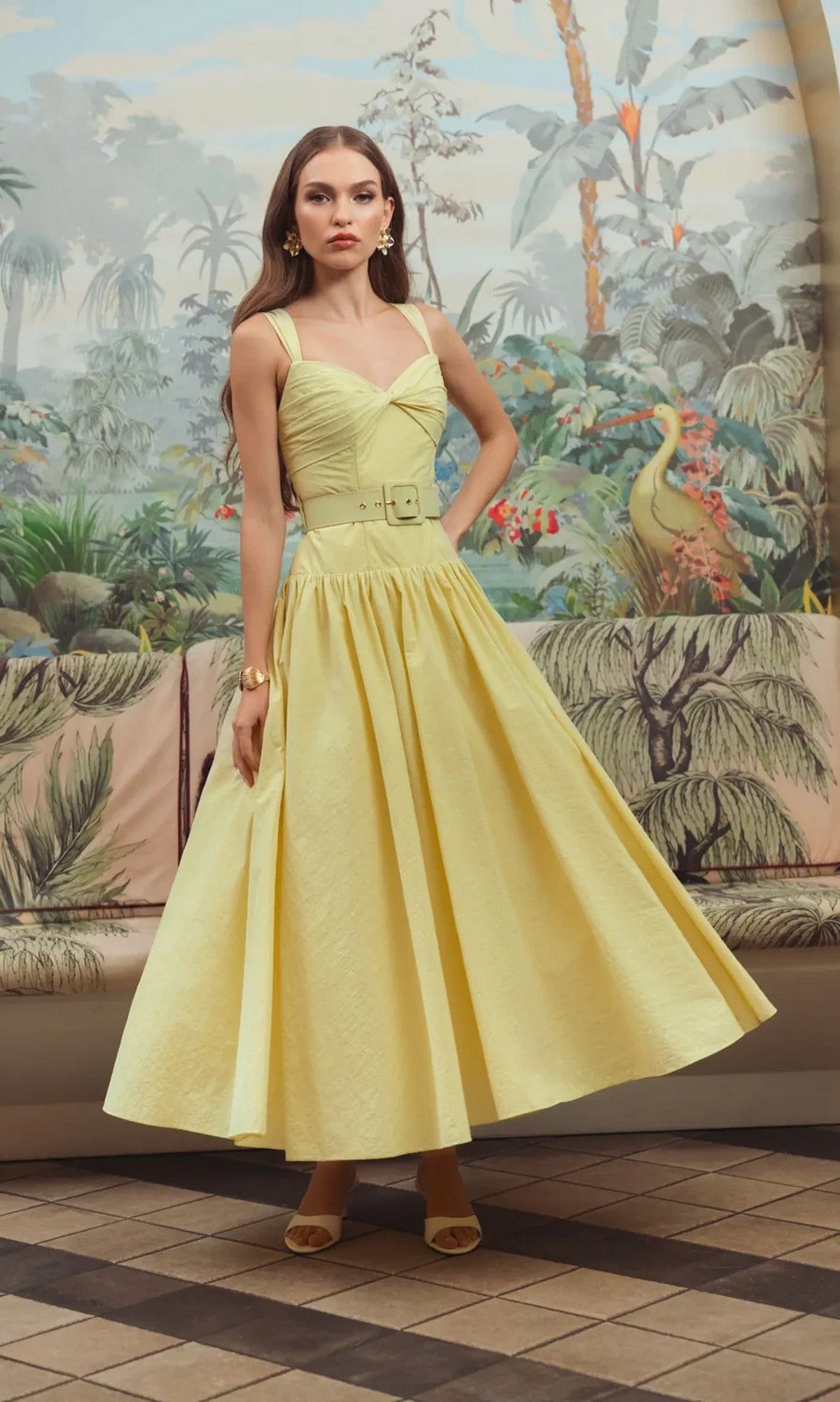 A woman in a buttercup Mac Duggal 21251 tea-length party dress and matching shoes stands indoors in front of a tropical-themed mural and patterned sofa. The vibrant dress adds a stylish touch to the lively setting.