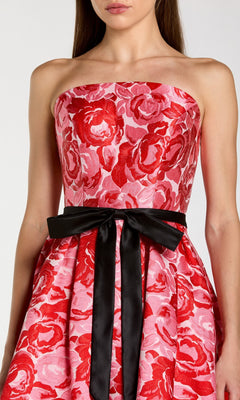 A woman is shown from shoulders to knees wearing the Tea-Length Party Dress: Mac Duggal 21252—a strapless pink dress featuring a bold red floral pattern and a black ribbon cinched at the waist.