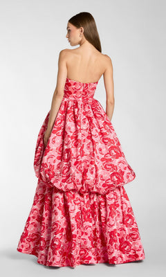 A woman stands facing away, wearing the Long Formal Dress: Mac Duggal 21254—a strapless pink floral ballgown with a voluminous skirt adorned with delicate red blooms.