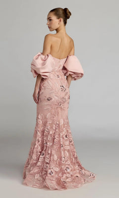 A woman wearing the Mac Duggal 21260 long formal dress—an off-the-shoulder blush pink satin trumpet gown with floral lace embroidery and puff sleeves—stands against a neutral background.