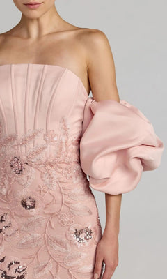 A woman wearing the Mac Duggal 21260 long formal dress—a strapless pink gown featuring intricate floral lace embroidery and a dramatic off-the-shoulder puff sleeve on her left arm.