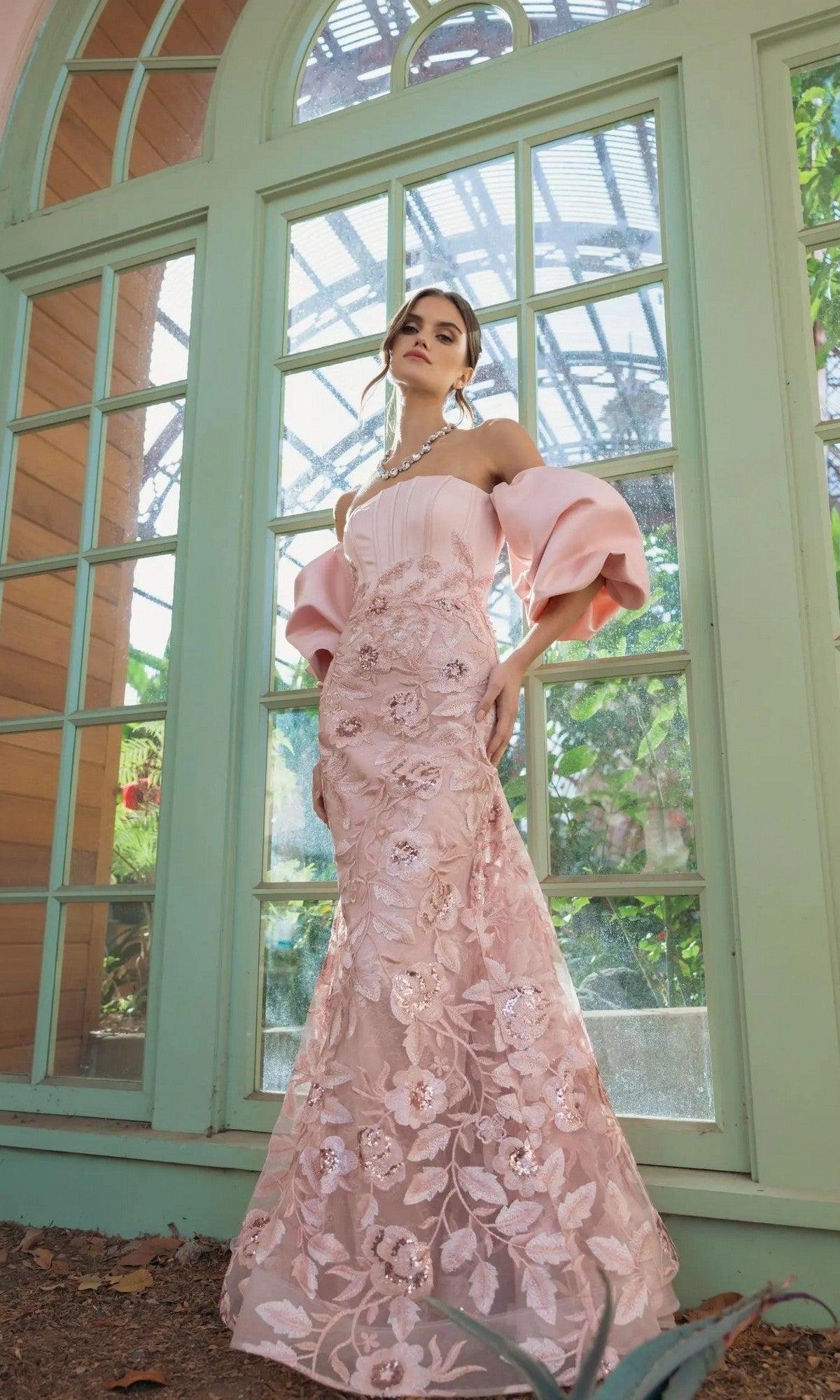 A woman wearing the Mac Duggal 21260 long formal dress—a pale pink, off-the-shoulder gown with puff sleeves and a floral design—stands in front of large arched windows with greenery visible outside.