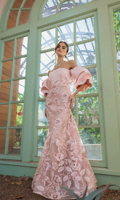 A woman wearing the Mac Duggal 21260 long formal dress—a pale pink, off-the-shoulder gown with puff sleeves and a floral design—stands in front of large arched windows with greenery visible outside.