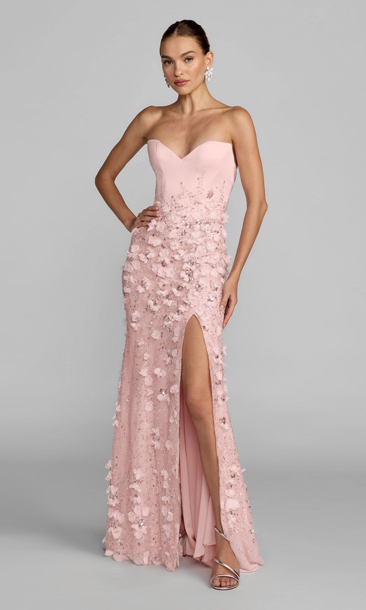 A woman wearing the Long Formal Dress: Mac Duggal 21261 in blush—a strapless gown with a sweetheart neckline, thigh-high slit, and delicate 3D flower embellishments—paired with silver heels, stands against a plain gray background.