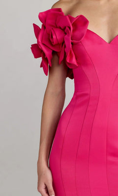 A person wearing the Mac Duggal 21263 Long Formal Dress—a fitted, vibrant pink gown featuring a dramatic, ruffled floral detail on one shoulder—shown from the upper chest to the hip.