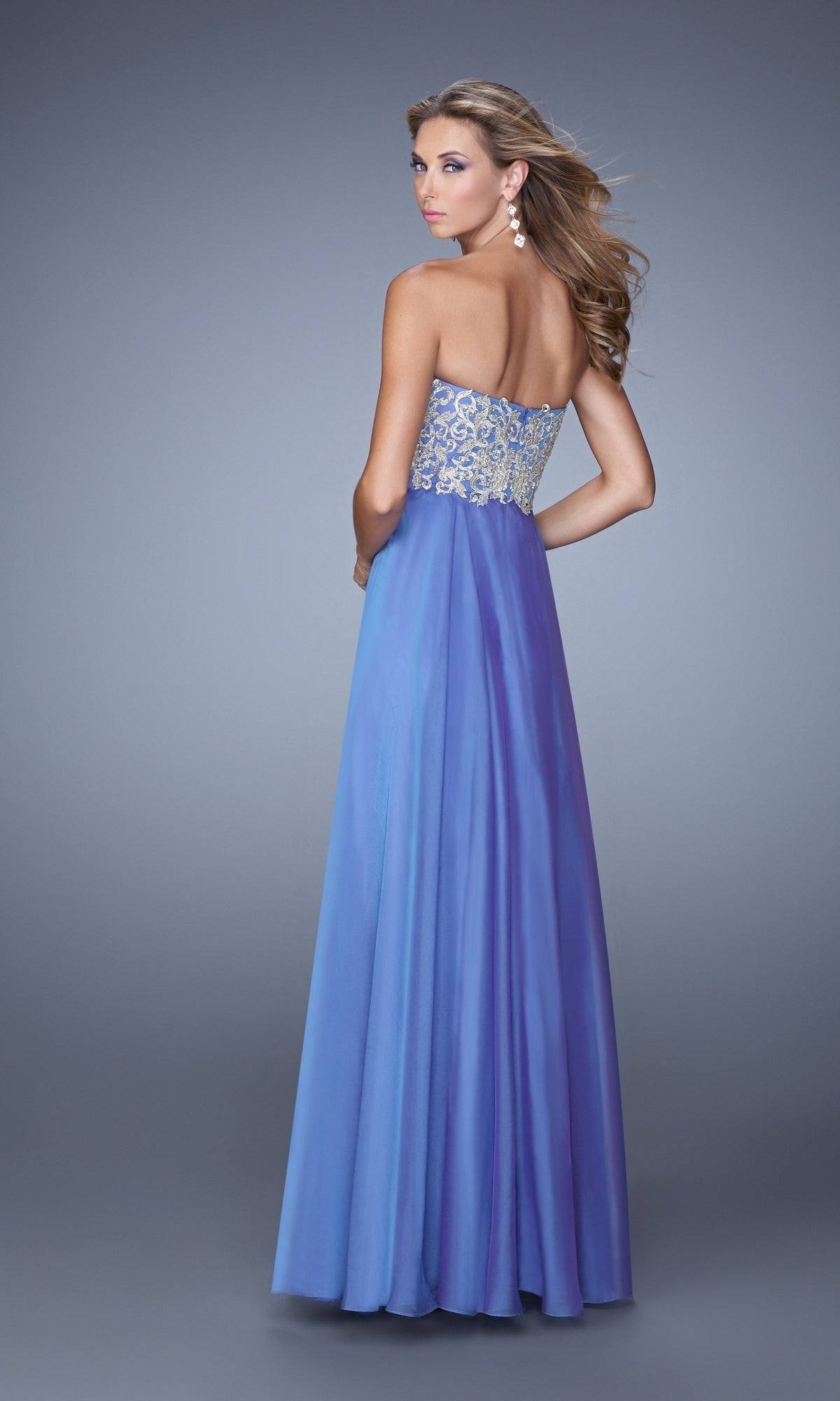 A woman faces away, wearing the La Femme Embroidered Chiffon Prom Dress 21289 in lavender with a beaded bodice and large earrings, set against a plain gray background.