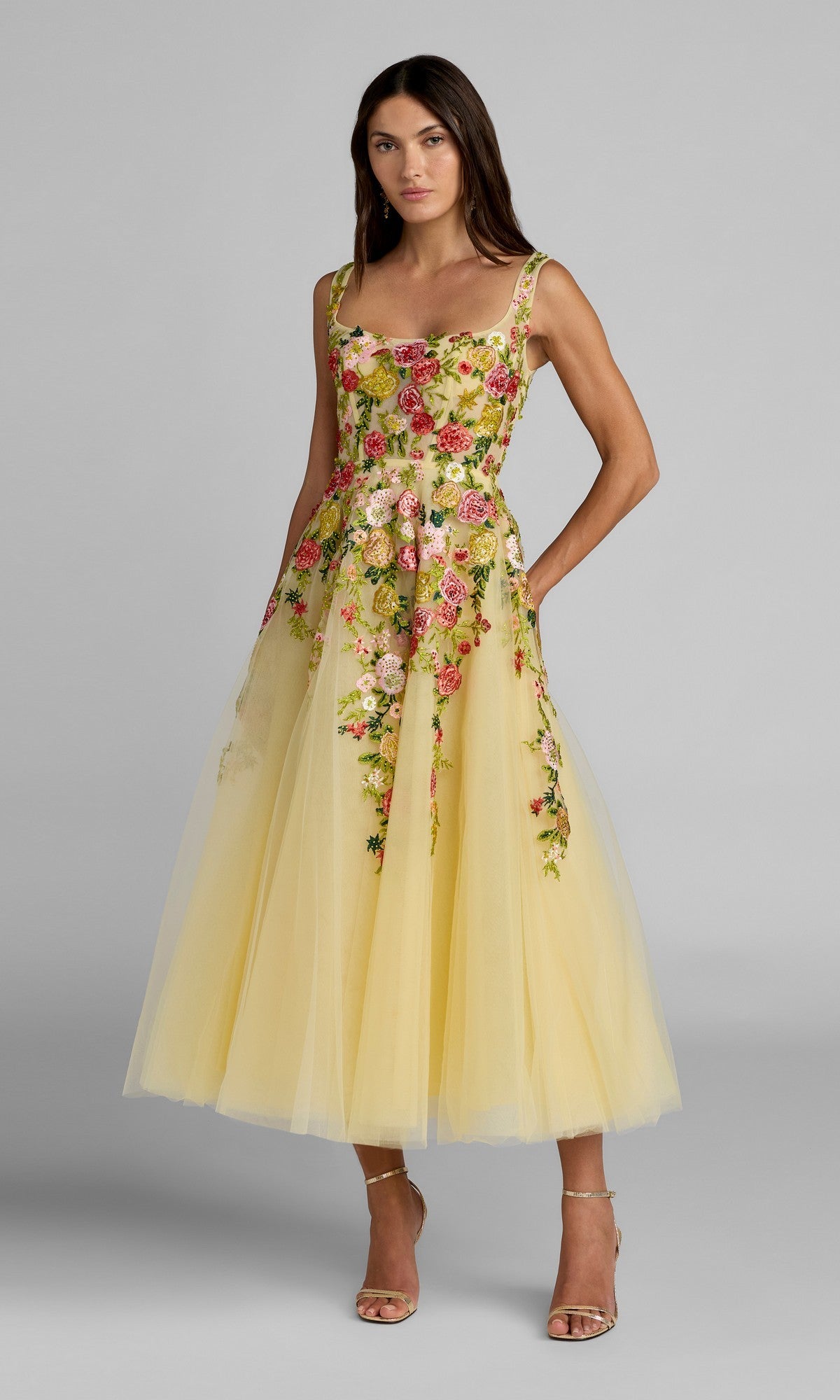 A woman stands against a plain gray background wearing the Mac Duggal 21294 Tea-Length Party Dress in Butter Multi, featuring floral embroidery, a square neckline, and bustier bodice. She pairs the look with nude strappy heels and maintains a neutral expression.