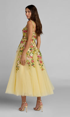 A woman stands in profile against a plain gray background, looking over her shoulder while wearing the Tea-Length Party Dress: Mac Duggal 21294—a sleeveless dress with a bustier bodice and square neckline, featuring ethereal yellow tulle and vibrant floral embroidery for a feminine look.