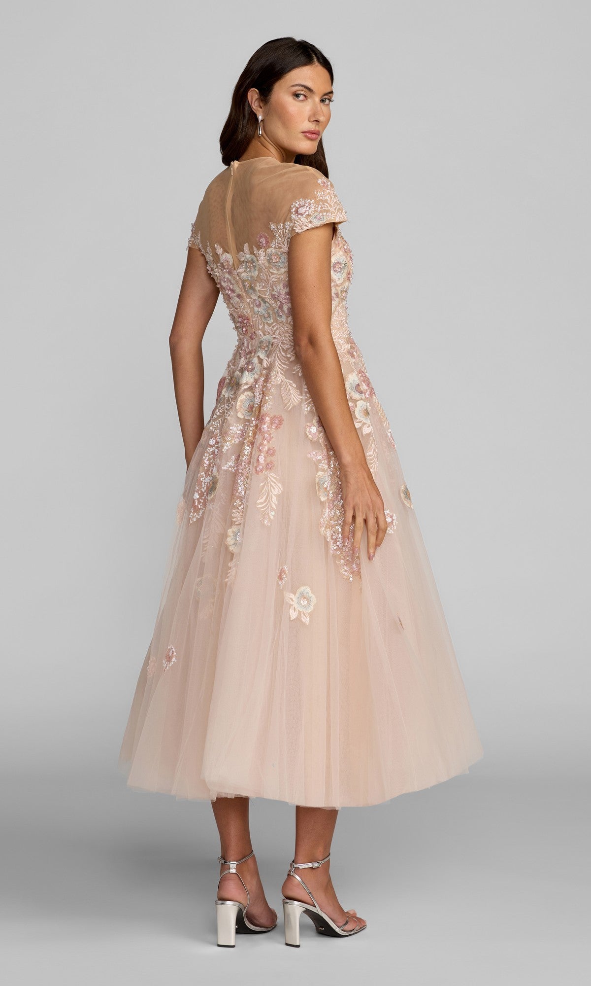 A woman stands facing slightly away, wearing the Mac Duggal 21296 Tea-Length Party Dress—a pink, short-sleeved floral embroidered dress featuring an a-line tulle skirt that falls to her ankles—paired with silver high-heeled sandals.