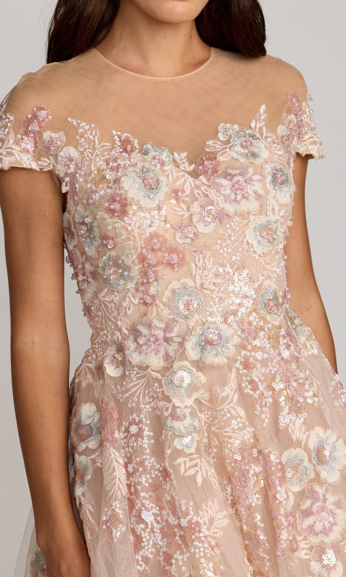 A woman wearing the Mac Duggal 21296 tea-length party dress, which features a sheer short-sleeved bodice embellished with pink and blue floral embroidery and sparkling sequin details, complemented by an elegant A-line tulle skirt.