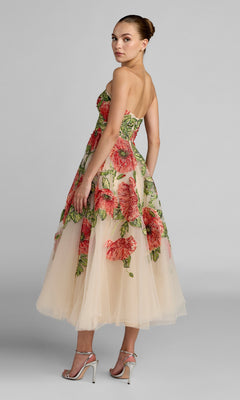 A woman stands sideways, looking over her shoulder, dressed in the Tea-Length Party Dress: Mac Duggal 21300—a strapless tulle midi dress adorned with bold red floral embroidery. She completes the sophisticated look with silver high-heeled sandals, all set against a gray background.