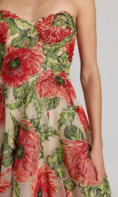 A person wearing the Mac Duggal 21300 tea-length party dress, featuring a strapless tulle design with large red and green floral embroidery, stands against a plain light background.