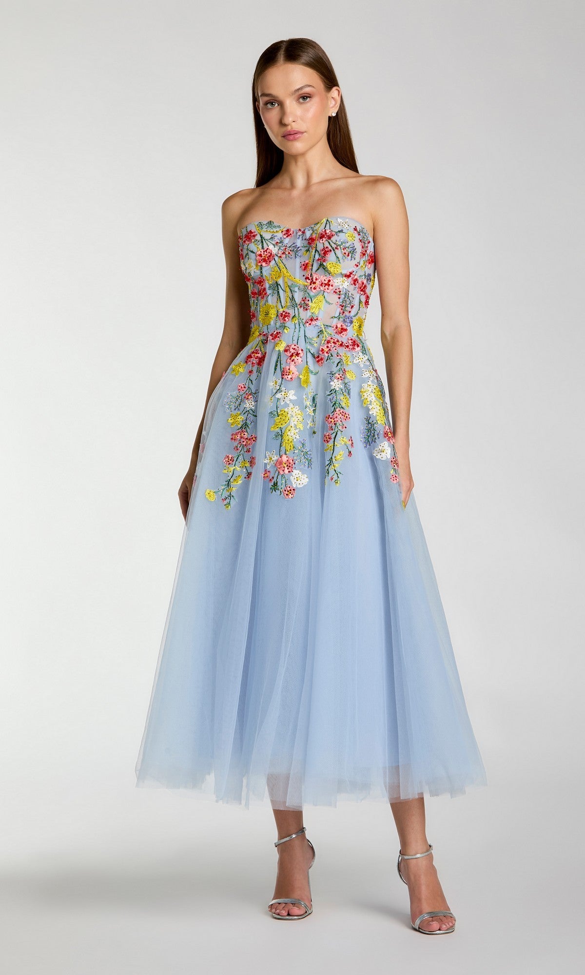 A woman stands against a plain light gray background, wearing the Tea-Length Party Dress: Mac Duggal 21304—a stunning strapless tea-length dress in Periwinkle Multi, crafted from light blue tulle and adorned with colorful floral embroidery.