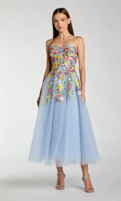 A woman stands against a plain light gray background, wearing the Tea-Length Party Dress: Mac Duggal 21304—a stunning strapless tea-length dress in Periwinkle Multi, crafted from light blue tulle and adorned with colorful floral embroidery.