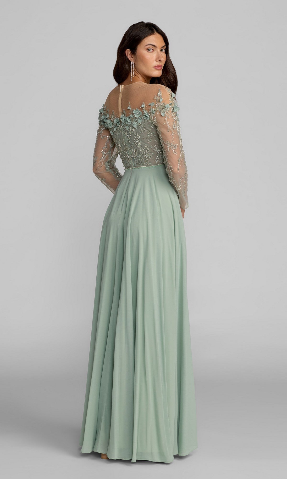 A woman wearing the Mac Duggal 21315 long formal dress in sage green, featuring sheer embroidered long sleeves and floral details on the bodice, stands against a plain gray background.