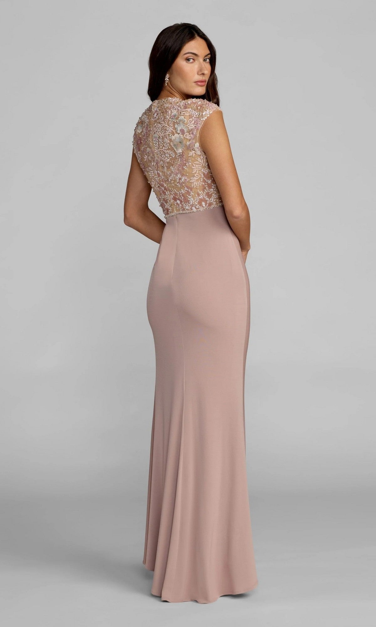 A woman stands against a plain gray background, looking over her shoulder in the Mac Duggal 21324 long formal dress—a floor-length, light mauve gown featuring a fitted, embroidered lace bodice and a column jersey skirt.
