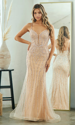 A woman in a fitted nude Long Beaded Mermaid Prom Gown, Amelia Couture 2132, stands before a mirror showing the gown’s front and back. With long, wavy hair, she is posed beside a vase of dried grasses.