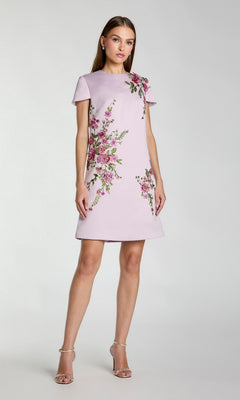 A woman wearing a short-sleeve, lilac Mac Duggal 21338 dress with floral embroidery stands against a plain light background and looks at the camera.