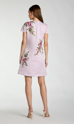 A woman stands facing away, wearing the Mac Duggal 21338 short wedding guest dress—a light pink, short-sleeved design accented with delicate floral embroidery—paired with beige high-heeled sandals against a plain background.