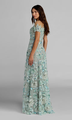A woman stands sideways against a plain gray background wearing the Mac Duggal 21339, a light blue off-shoulder, floor-length formal gown adorned with delicate beaded floral embroidery.