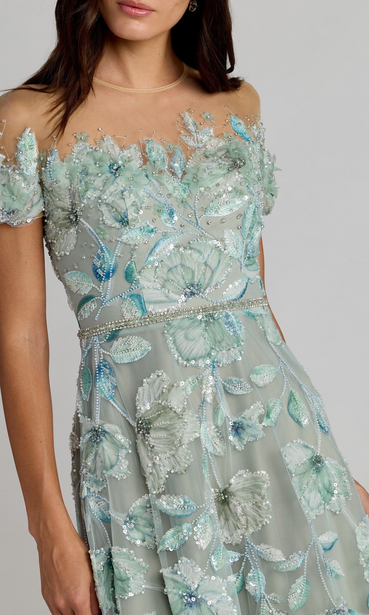 A woman wearing the Mac Duggal 21339 long formal dress in sage green, featuring an off-shoulder sheer design, blue-green beaded floral embroidery, and exquisite bead and sequin detailing.