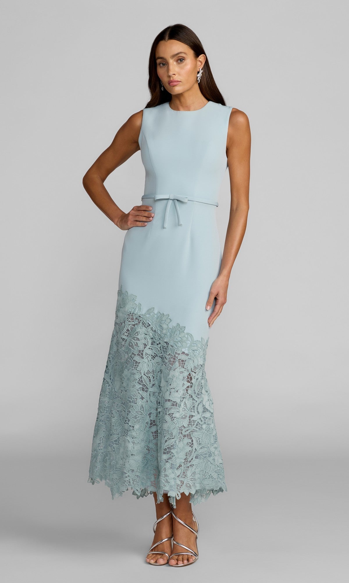A woman stands in sandals against a plain gray background, wearing the Tea-Length Party Dress: Mac Duggal 21355 in powder blue—a sleeveless crepe dress adorned with delicate lace detailing at the hem and an asymmetrical lace skirt.