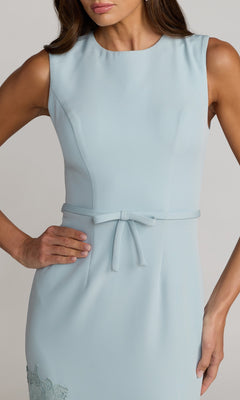 A woman models the Mac Duggal 21355 Tea-Length Party Dress, a sleeveless light blue crepe design with a thin belted bow at the waist and an elegant asymmetrical lace skirt, posing with one hand on her hip.
