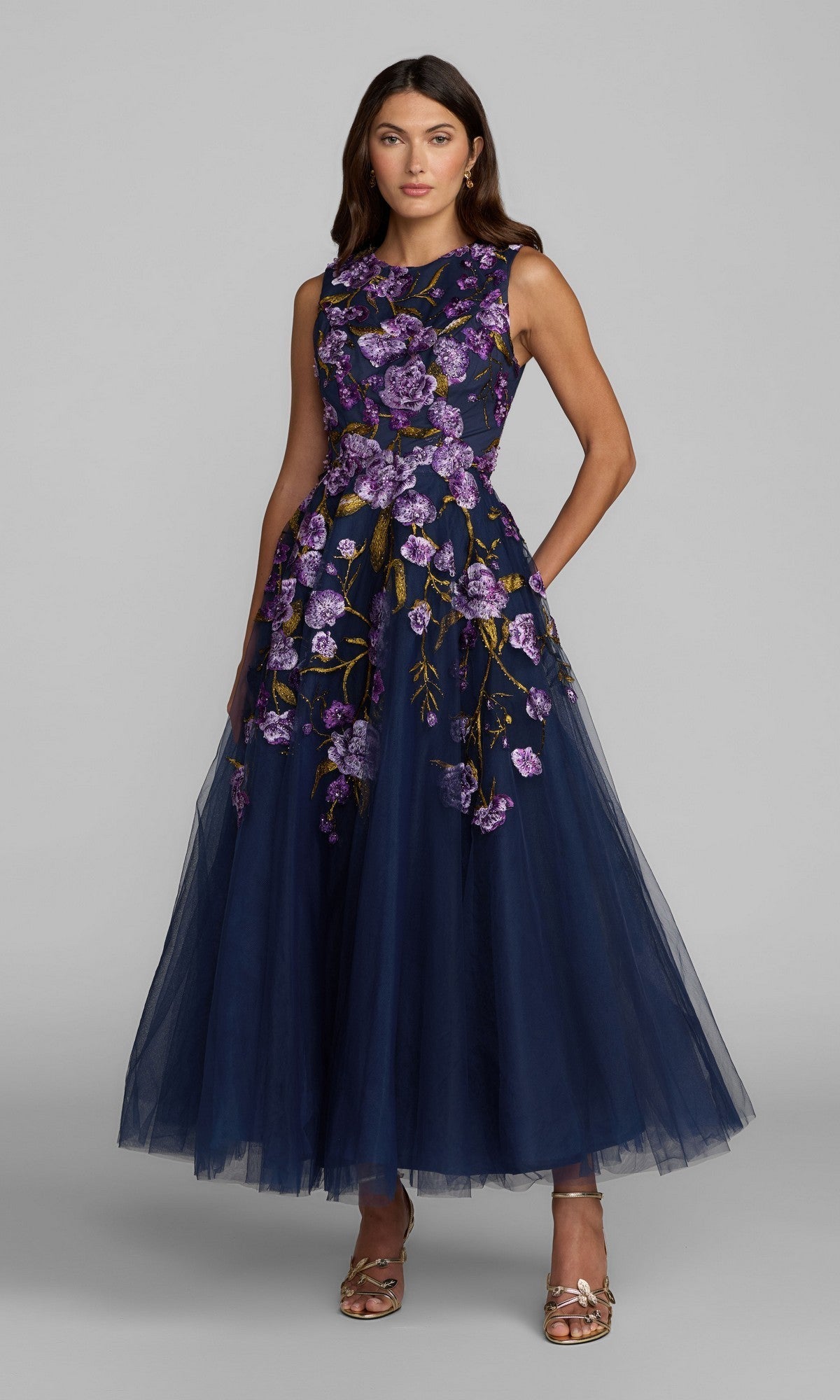 A woman wears the ankle-length Mac Duggal 21358 party dress in Midnight Multi, featuring a sleeveless design with purple floral embroidery and a tulle A-line skirt, paired with gold sandals, as she stands against a plain gray background.