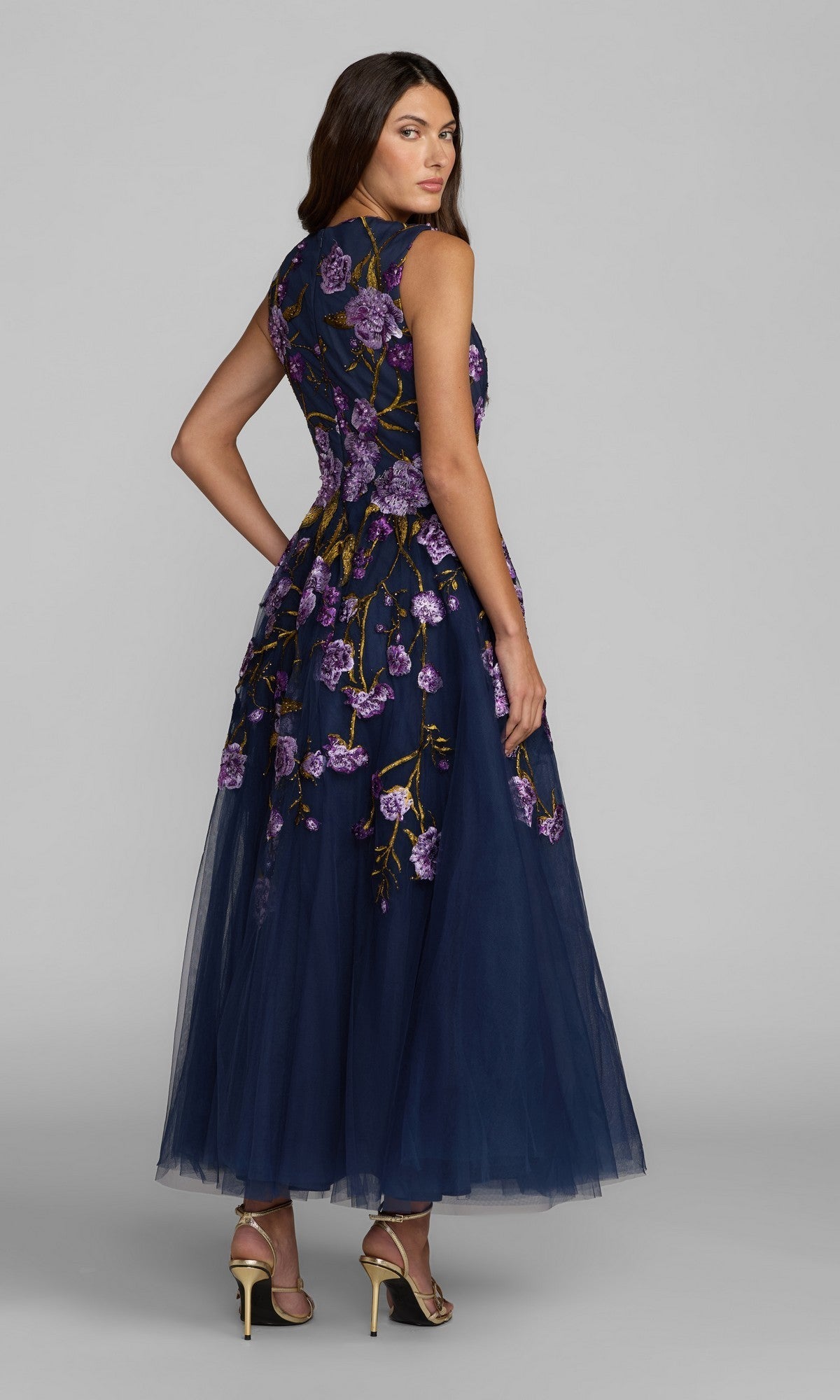 A woman stands against a gray background in the Mac Duggal 21358 ankle-length party dress, featuring sleeveless navy blue floral embroidery and a tulle A-line skirt. She wears heeled sandals and looks over her shoulder.