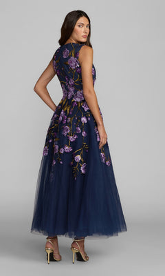 A woman stands against a gray background in the Mac Duggal 21358 ankle-length party dress, featuring sleeveless navy blue floral embroidery and a tulle A-line skirt. She wears heeled sandals and looks over her shoulder.