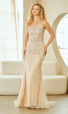 A woman stands indoors in front of a white sofa and arched window, wearing the Amelia Couture 2136-2134 Lace-Up Beaded Prom Gown—a floor-length, fitted, sleeveless beige dress with a corset-style bodice and silver bead detailing.