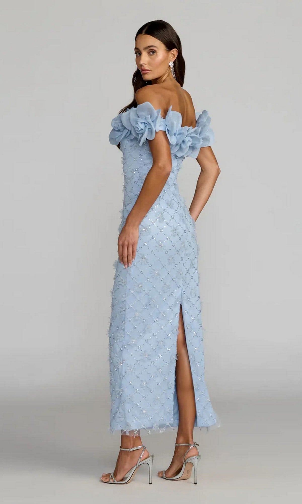 A woman wearing the Mac Duggal 21363 Midi Party Dress, a light blue, off-the-shoulder, mid-length dress adorned with floral embellishments and featuring a back slit, paired with silver high heels, stands against a plain background.