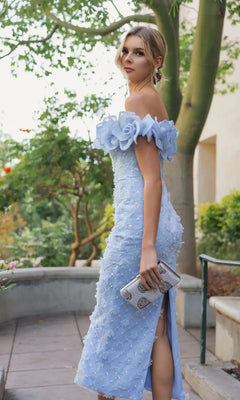 A woman wearing the Mac Duggal 21363 midi party dress in light blue, featuring an off-the-shoulder neckline and delicate floral accents, holds a clutch as she stands outdoors surrounded by trees and greenery.