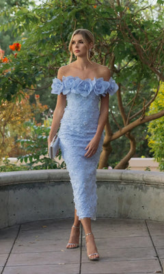 A woman stands outdoors wearing the Mac Duggal 21363 midi party dress in light blue, which features an off-the-shoulder neckline adorned with large floral appliqués and delicate small embellishments. She completes the look with a clutch bag and strappy heels.
