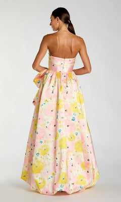 A woman stands with her back to the camera against a plain background, wearing the Mac Duggal 21376 Long Formal Dress—a strapless Mikado ballgown that sweeps the floor and features a vivid pink, yellow, and blue floral pattern. An oversized bow at the waist elevates the look with a striking touch of elegance.