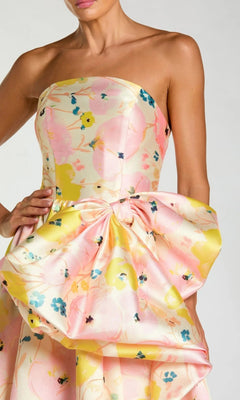 A woman wearing the Mac Duggal 21376 long formal dress, a strapless Mikado ballgown adorned with a pastel floral pattern and accented by an oversized bow at the waist.