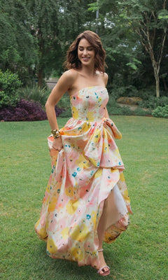 A woman wearing the Mac Duggal 21376 Long Formal Dress in Pink Multi, a strapless Mikado ballgown with a pastel floral design and an oversized bow at the waist, walks on grass in a garden surrounded by trees and shrubs.