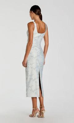 A woman is standing with her back to the camera against a plain background, wearing the Mac Duggal 21377 Midi Party Dress in light blue brocade featuring a back slit, paired with high-heeled sandals.