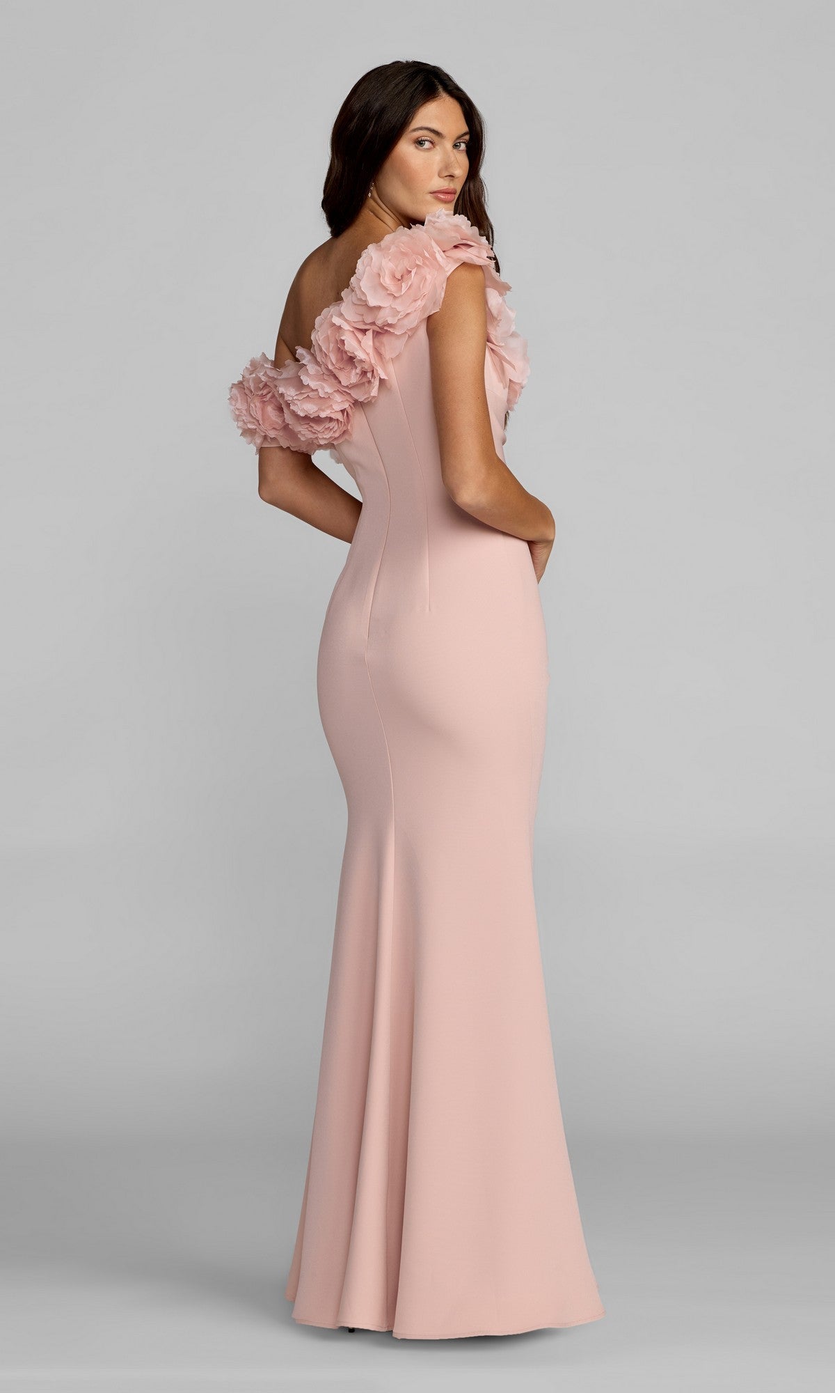 Woman in a light pink, floor-length Mac Duggal 21383 formal dress with striking 3D floral appliques on the shoulder, standing and facing backward with her head turned toward the camera.