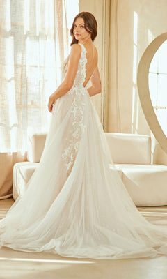A woman stands indoors in the Amelia Couture 2140, a white, floor-length wedding dress with tulle, lace embroidery, sheer details, and a corset bodice as she faces slightly away from the camera.