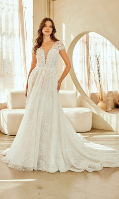 A woman stands indoors wearing the Amelia Couture 2142 long wedding dress with a lace A-line silhouette, corset bodice, cap sleeves, and a long train. Neutral furniture and sheer curtains are visible in the background.